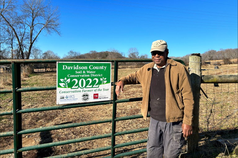 A man in a brown jacket stands next to a fence with a sign that reads "Davidson County Soil & Water Conservation District."