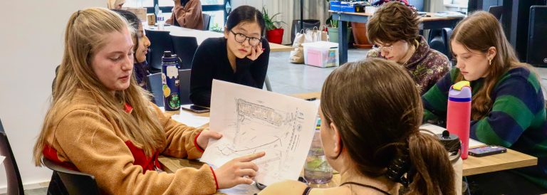 A female student reviews a diagram with other students seated around a work table.