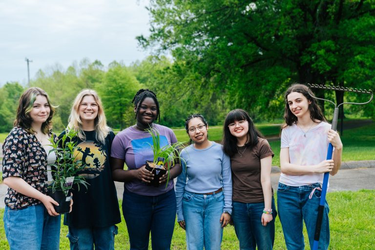 Six teenagers face the camera holding plants in their garden.