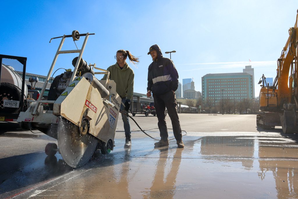 Two men use heavy machinery to cut through concrete pavement.
